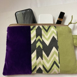 May include: A rectangular clutch with a gold zipper, featuring a purple velvet panel, a chevron-patterned panel in black, green, and cream, and a green canvas panel. The clutch contains a smartphone, a white charging case, and a lipstick.