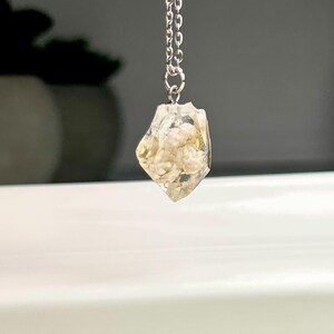 May include: A silver chain necklace with a clear, geometric pendant. The pendant is filled with a light beige, textured material. The necklace is displayed against a blurred background.