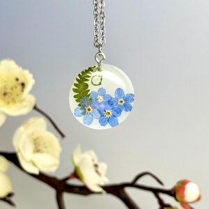 May include: A silver chain necklace with a round pendant featuring a pressed forget-me-not flower design in a clear resin setting.