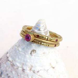Pode incluir: Um conjunto de dois anéis dourados com superfície texturizada e pequenas pedras redondas de cor magenta. Os anéis estão empilhados, um com uma pedra magenta maior engastada, enquanto o outro possui pedras magenta menores incrustadas ao longo da banda.
