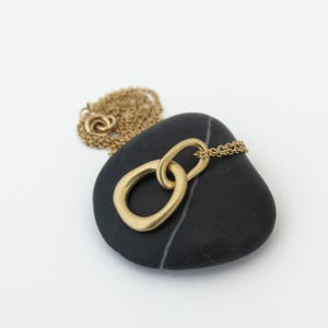 May include: A gold necklace with a simple, minimalist pendant featuring two interlocking circles. The pendant is resting on a black stone.