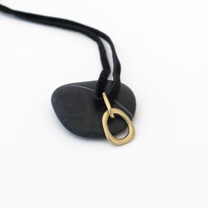 May include: A gold oval pendant necklace with a simple, organic shape. The necklace is on a black suede cord and is resting on a black stone.