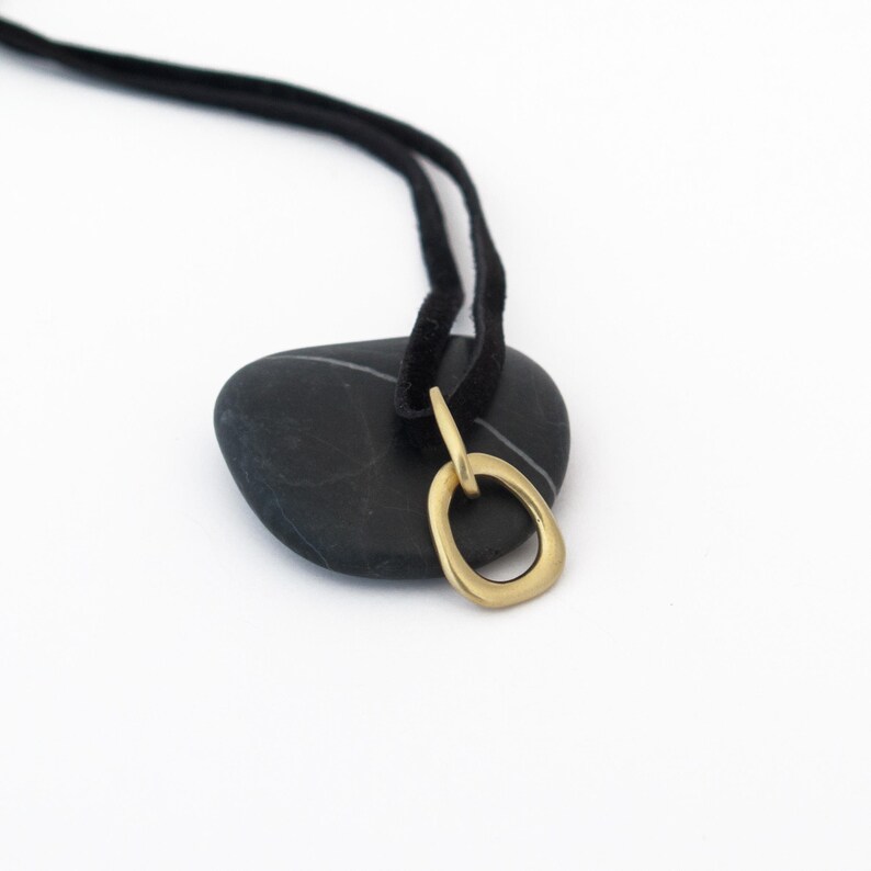 May include: A gold oval pendant necklace with a simple, organic shape. The necklace is on a black suede cord and is resting on a black stone.
