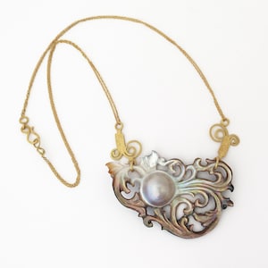 Peut inclure: Un collier en chaîne dorée avec un grand pendentif orné en nacre irisée. Le pendentif présente une grande perle ronde au centre et des motifs complexes et tourbillonnants.