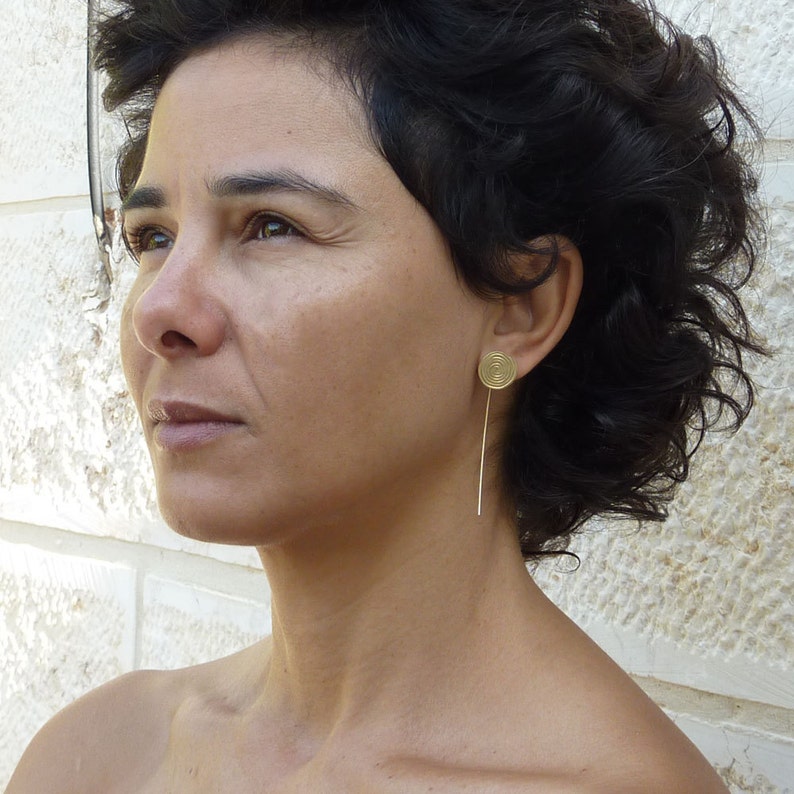 May include: A woman with short, dark curly hair wearing a pair of gold earrings with a spiral design. The earrings are long and dangle from the earlobes.