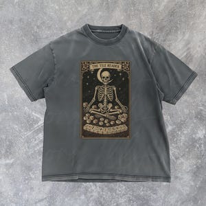 May include: Dark grey t-shirt featuring a tarot card design. The graphic depicts a skeleton meditating with tiles, a crescent moon, and the text "THE TILE READER". The design is in shades of brown and cream.