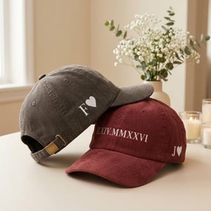 Puede incluir: Una gorra de béisbol de pana gris carbón con una letra F blanca y un icono de corazón, y una gorra de pana granate con el texto en números romanos II.XIV.MMXXVI y la letra J con un icono de corazón, ambas con hebillas de tono latón.
