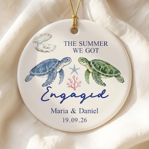 May include: A round white ceramic ornament featuring a blue sea turtle and a green sea turtle, a small starfish, pink coral, and an open oyster shell with a pearl. The text reads THE SUMMER WE GOT Engaged Maria & Daniel 19.09.26 in blue font.