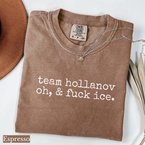 May include: Espresso-colored t-shirt with the text "team hollanov oh, & fuck ice." A silver necklace with a small pendant rests on the shirt. The shirt has a Comfort Colors tag.
