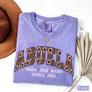 May include: A violet Comfort Colors t-shirt with the word "ABUELA" in a brown floral design. Below the word are the names Maria, Juan, Mario, Byanca, and Jose. A gold necklace and a brown hat are also in the image.