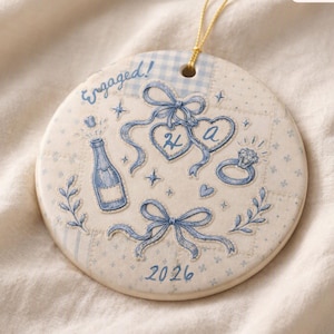 Puede incluir: Un adorno de cerámica redondo con fondo crema y patrones de estilo patchwork en azul. Incluye el texto Engaged y 2026, con ilustraciones de una botella de champán, dos etiquetas en forma de corazón con H y A, un anillo de diamantes y lazos.