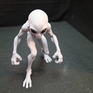 May include: A light grey alien figurine in a crouching, predatory stance. The creature features a smooth, elongated head, large dark oval eyes, and slender limbs with defined musculature. Its hands and feet have elongated, distinct digits.