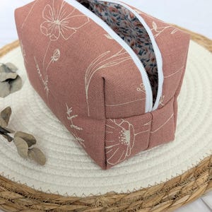 Könnte beinhalten: Ein rechteckiges Reißverschlussetui in Altrosa mit einem weißen floralen Linienmuster. Das Etui verfügt über einen weißen Reißverschluss und ein Innenfutter mit einem kleinen blau-roten Blumenmuster auf weißem Hintergrund.