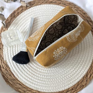 Könnte beinhalten: Senfgelbe Kosmetiktasche mit weißer Blumenstickerei, geöffnet, um ein braunes Blumenmuster im Inneren zu zeigen. Die Tasche liegt auf einer gewebten Matte mit einem Make-up-Pinsel und einem kleinen Glasbehälter.