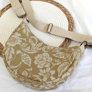 Könnte beinhalten: Eine halbmondförmige Umhängetasche mit beigem Blumenmuster auf tanfarbenem Hintergrund. Die Tasche hat einen passenden verstellbaren beigen Riemen und bronzefarbene Hardware. Die Tasche liegt auf einer weißen Oberfläche mit einer gewebten Strohmatte.
