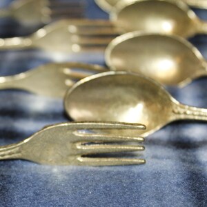 May include: A set of five gold-toned metal spoons and forks with ornate handles. The spoons and forks are arranged in a row, with the handles pointing to the right.