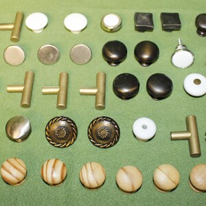 May include: A collection of various cabinet knobs and T-bar pulls in assorted materials and finishes, including brushed brass, dark wood, white ceramic, matte black, and metallic textures, arranged in rows on a flat green surface.
