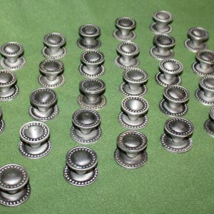 May include: A collection of 36 silver-toned cabinet knobs with a beaded rim and a raised center. The knobs are arranged in rows on a green surface.