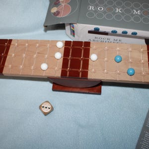 May include: A wooden board game with a light brown and dark brown design. The game includes white and blue spheres, a die, and a box with the game's name, "Rock Me Archimedes".