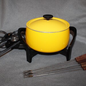 May include: A yellow fondue pot with a black base and lid, and a set of fondue forks. The pot has a black handle and a black knob on the lid. The forks have wooden handles.