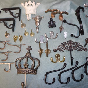 May include: A collection of various metal wall hooks and brackets in assorted finishes including black, brass, silver, and white. The assortment features crown-shaped hooks, ornate scrollwork designs, simple double-pronged hooks, and industrial-style brackets.