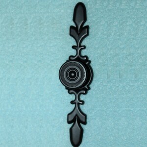 May include: A black, ornate cabinet pull with a circular center and decorative, symmetrical extensions. The pull is made of metal and features a detailed, textured design. The background is a light blue color.