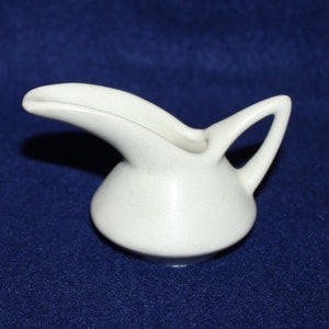 May include: A small, white ceramic creamer with a curved spout and a handle. The creamer has a smooth, matte finish.