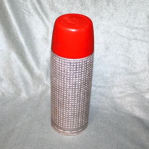 May include: A vintage thermos with a bright red lid and a silver, perforated body. The thermos is cylindrical and appears to be made of metal. The lid has a small, embossed design. The thermos is designed to keep liquids hot or cold.