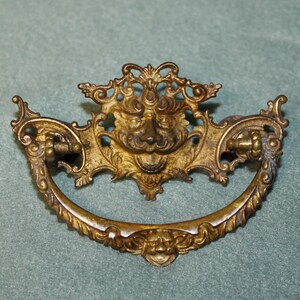 May include: A brass-toned metal drawer pull featuring an ornate, symmetrical design with a central lion head motif. The hardware includes a curved, swinging handle with decorative scrollwork and a smaller face detail at the bottom center point.