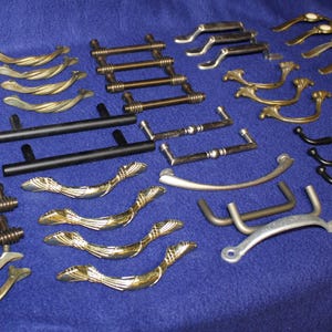 May include: A collection of various metal cabinet hardware pulls and handles in assorted finishes including polished gold, brushed bronze, matte black, and silver. The assortment features straight bars, curved designs, and textured detailing.