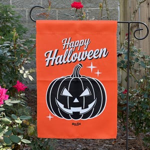 Op de afbeelding: Een rechthoekige oranje tuin vlag met de tekst Happy Halloween in het wit met een zwarte omlijning. Onder de tekst staat een grote zwarte pompoen met witte ogen, neus en mond, geaccentueerd door twee kleine witte vierpuntige sterren.