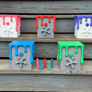 Può includere: Sei piccole casseforti in metallo grigio con vernice di diversi colori che cola sui lati. I colori sono rosa, rosso, viola, blu, verde e una sesta cassaforte senza vernice. Le casseforti sono disposte in fila su una superficie di legno.