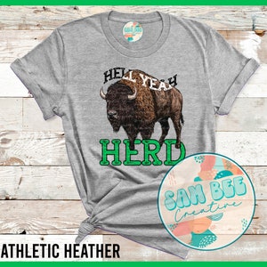 May include: A gray t-shirt with a green and white graphic of a buffalo and the text "Hell Yeah Herd". The shirt is on a wooden surface. The text "Sam Bee Creative" is on a circle with a colorful abstract design.