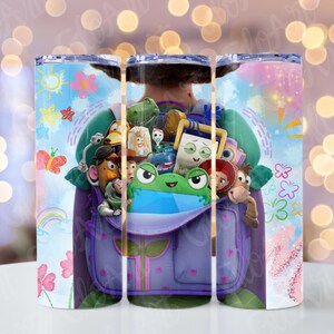 May include: A cylindrical tumbler featuring a colourful graphic print of a purple backpack filled with various cartoon characters, including a green frog, a cowboy, a space ranger, and a potato figure, surrounded by drawings of flowers and a rainbow.
