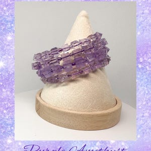 May include: A purple amethyst gemstone bracelet with multiple strands of faceted beads. The bracelet is displayed on a wooden stand.
