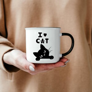 Pu&ograve; includere: Tazza in ceramica bianca con manico nero. La tazza ha un'illustrazione in bianco e nero di un gatto che sporge dal bordo e il testo "I &hearts; CAT" stampato sul davanti.