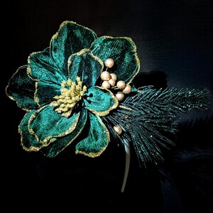 May include: A teal velvet flower with gold glitter edging and gold berries. A sprig of glittery green pine and a white headband complete the floral hair accessory. The background is black.
