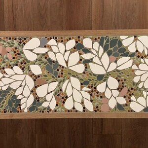 Può includere: Un piano da tavolo in mosaico con un design floreale. Il design presenta foglie e fiori bianchi su uno sfondo di piastrelle verdi, marroni e rosa.