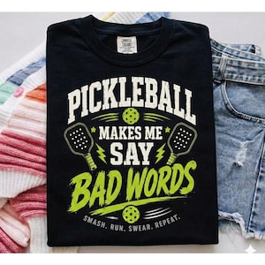 May include: Black t-shirt with the text "PICKLEBALL MAKES ME SAY BAD WORDS" in white and green. The design includes pickleball paddles and balls. The phrase "SMASH. RUN. SWEAR. REPEAT." is printed at the bottom.