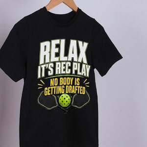 Könnte beinhalten: Schwarzes Kurzarm-T-Shirt mit dem Text "RELAX IT'S REC PLAY NO BODY IS GETTING DRAFTED" in Weiß und Gelb. Das Shirt zeigt eine Pickleball- und Schlägergrafik. Das Shirt hängt an einem Holzbügel.