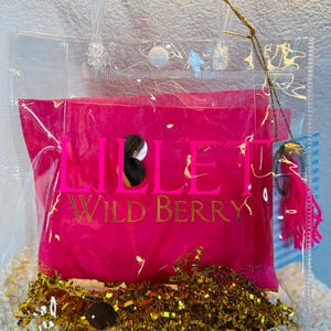 May include: A clear tote bag with a hot pink interior, featuring gold text that reads "LILLET WILD BERRY." The bag contains gold metallic filler and a small charm with pink tassels. The bag has clear handles and a snap closure.