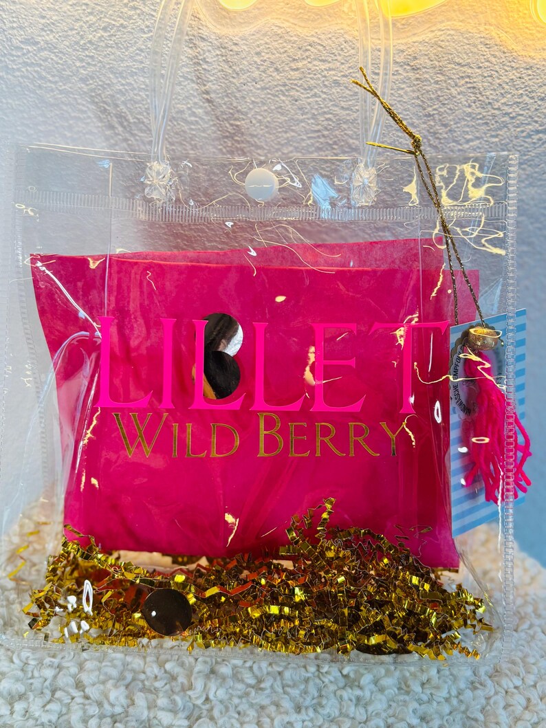 May include: A clear tote bag with a hot pink interior, featuring gold text that reads "LILLET WILD BERRY." The bag contains gold metallic filler and a small charm with pink tassels. The bag has clear handles and a snap closure.