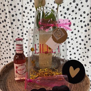 K&ouml;nnte beinhalten: Eine transparente Tragetasche mit dem Text "Lillet Wild Berry" enth&auml;lt wei&szlig;e Blumen und goldene Konfetti. Eine Flasche Lillet Berry, rosa herzf&ouml;rmige Sonnenbrillen und ein schwarzer Untersetzer mit goldenem Herz befinden sich in einem geflochtenen Korb.