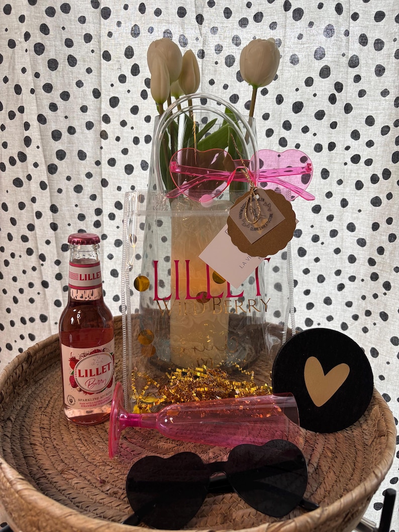 K&ouml;nnte beinhalten: Eine transparente Tragetasche mit dem Text "Lillet Wild Berry" enth&auml;lt wei&szlig;e Blumen und goldene Konfetti. Eine Flasche Lillet Berry, rosa herzf&ouml;rmige Sonnenbrillen und ein schwarzer Untersetzer mit goldenem Herz befinden sich in einem geflochtenen Korb.