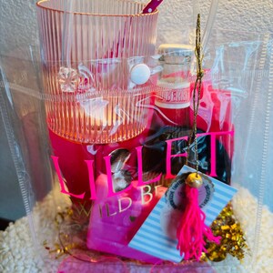 May include: A clear tote bag filled with pink items. Includes a tall, ribbed glass, a small bottle, a pink straw, sunglasses, and a tag with a pink tassel. The bag has a white button closure and the text "Lillet Wild Berry".