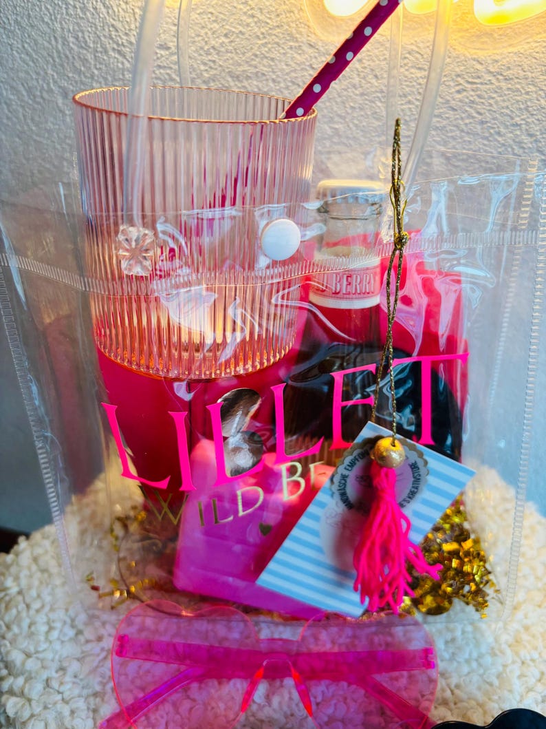 May include: A clear tote bag filled with pink items. Includes a tall, ribbed glass, a small bottle, a pink straw, sunglasses, and a tag with a pink tassel. The bag has a white button closure and the text "Lillet Wild Berry".