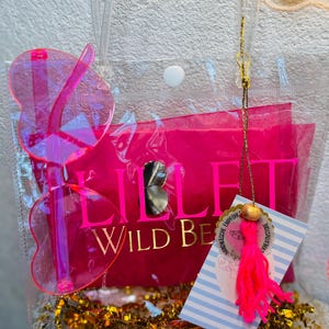 May include: A clear tote bag with a hot pink interior and butterfly-shaped sunglasses. The bag contains a tag with a pink tassel and gold accents. The text "LILLI WILD BE" is visible on the pink interior.