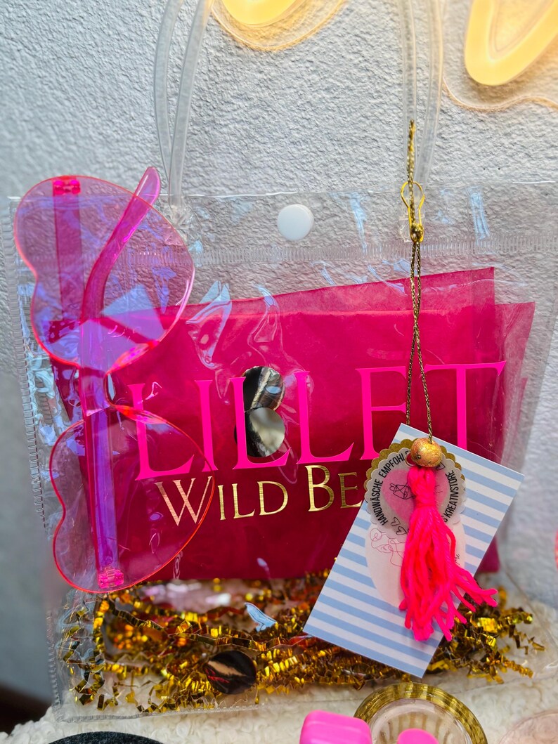 May include: A clear tote bag with a hot pink interior and butterfly-shaped sunglasses. The bag contains a tag with a pink tassel and gold accents. The text "LILLI WILD BE" is visible on the pink interior.