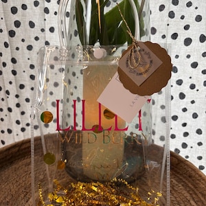 K&ouml;nnte beinhalten: Eine transparente Tragetasche mit dem Text "LILLI WILD BERRY" in Pink und Gold. Die Tasche enth&auml;lt goldenes Konfetti und ist mit goldenen kreisf&ouml;rmigen Akzenten verziert. Im Hintergrund ist eine Glasvase mit wei&szlig;en Blumen zu sehen.