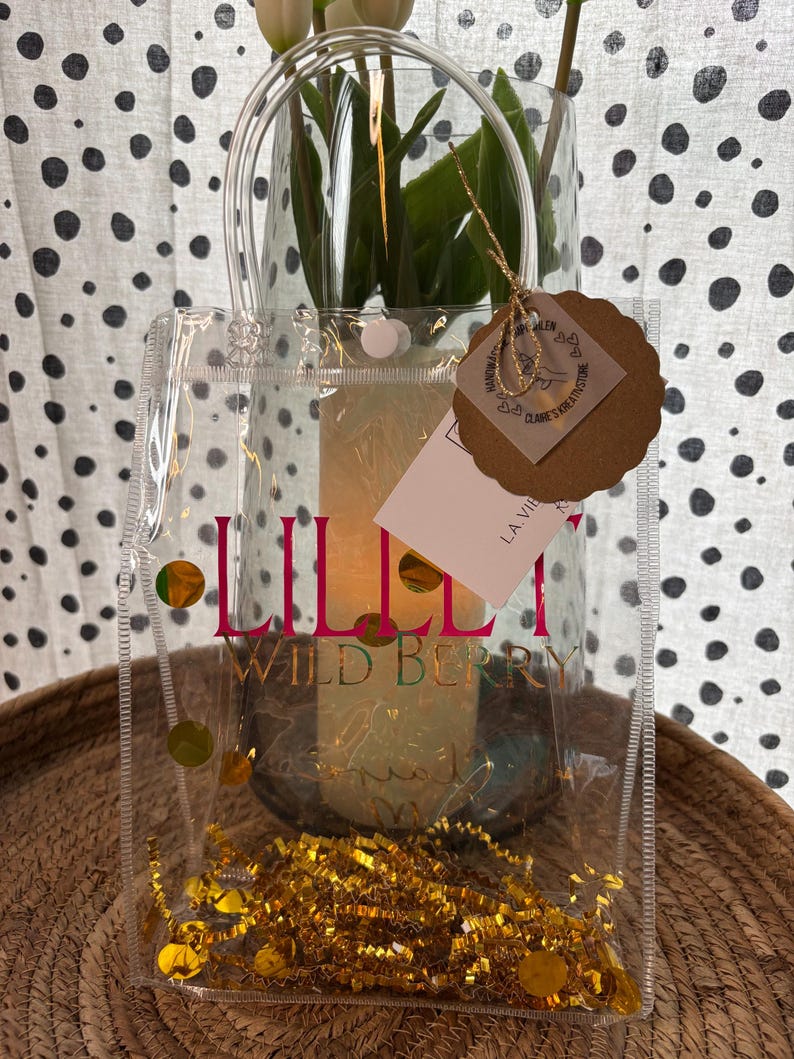 K&ouml;nnte beinhalten: Eine transparente Tragetasche mit dem Text "LILLI WILD BERRY" in Pink und Gold. Die Tasche enth&auml;lt goldenes Konfetti und ist mit goldenen kreisf&ouml;rmigen Akzenten verziert. Im Hintergrund ist eine Glasvase mit wei&szlig;en Blumen zu sehen.
